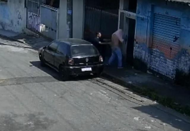 Homem sendo conduzido por outro, com a camisa levantada, para dentro de imóvel em rua, com carro estacionado, durante o dia, em cena de sequestro em cativeiro