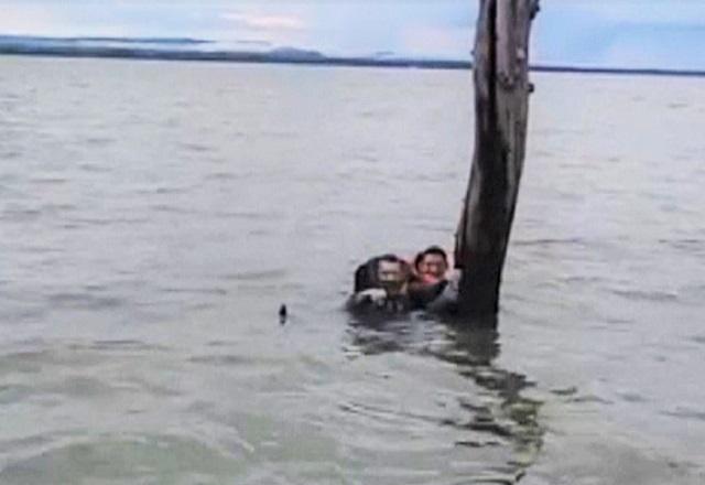 Homens de colete salva vidas agarrados em tronco fincado no meio de lago, com embarcação de resgate se aproximando, durante o dia