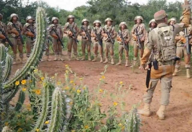 Homens fardados, empunhando armas, recebendo instruções de homem fardado em pé e com o braço levantado, ao lado de vegetação espinhosa, durante o dia, com céu azul em ambiente árido