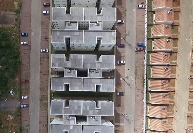 Imagem aérea de conjunto de prédios residenciais ao centro, à esquerda região de mata e, à direita, conjunto de casas com a visão dos telhados