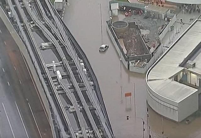 Imagem aérea de região com trilho de trem e avenida alagados, durante o dia, com carro submerso