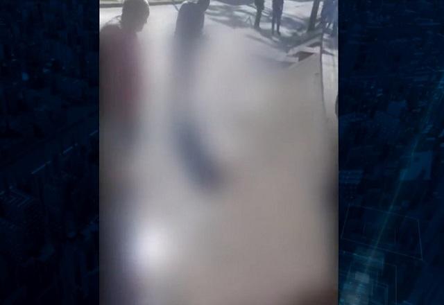 Imagem desfocada de pessoa morta no asfalto, com pessoas ao redor observando, durante o dia
