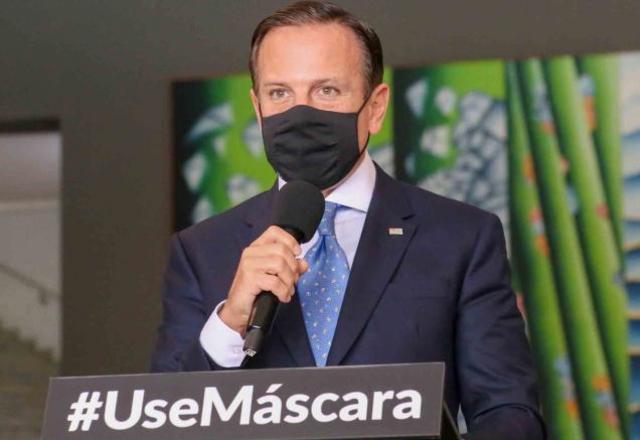 Imagem do governador João Doria. Ele está de terno, usa uma máscara de proteção da cor preta e segura um microfone
