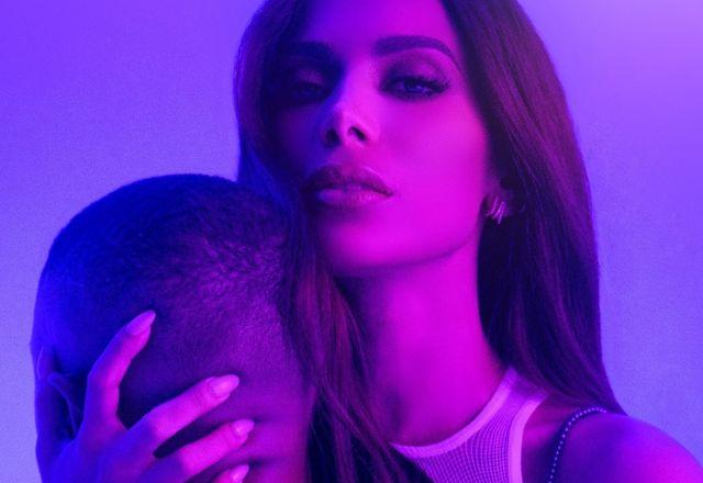 Imagem em todos de roxo que contém um casal abraçado. A mulher, que é Anitta, olha para a câmera