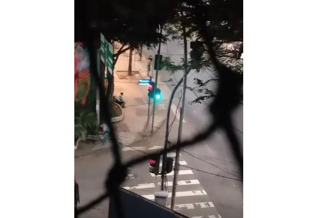 Imagem feita através de tela de janela, um homem armado numa esquina e outro se arrastando na calçada