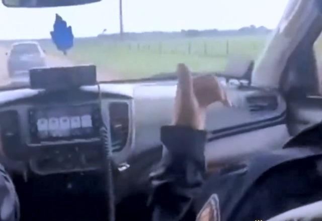 Imagem filmada de dentro de veículo, com passageira no banco da frente com farda e, à frente, carro sendo perseguido em estrada