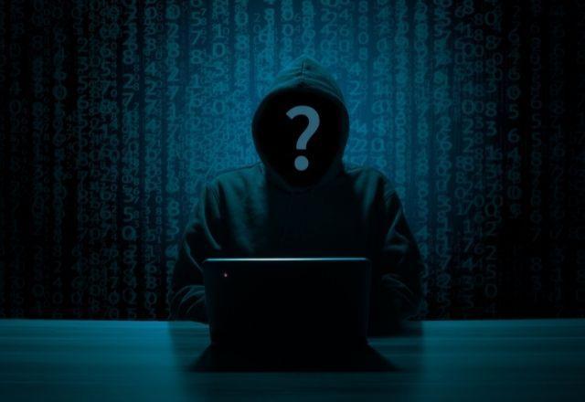 Imagem mostra, de forma sombria, um hacker em frente à tela de um notebook. No lugar do rosto, há um ponto de interrogação