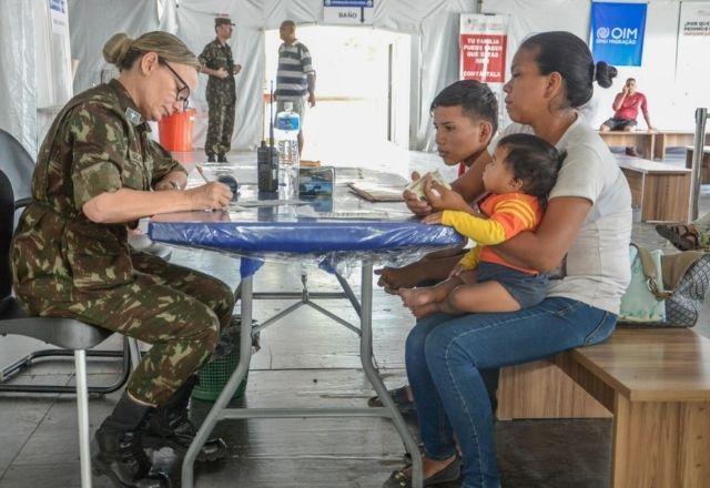 Imagem mostra família sentada à mesa na frente de uma militar que atende os imgrantes