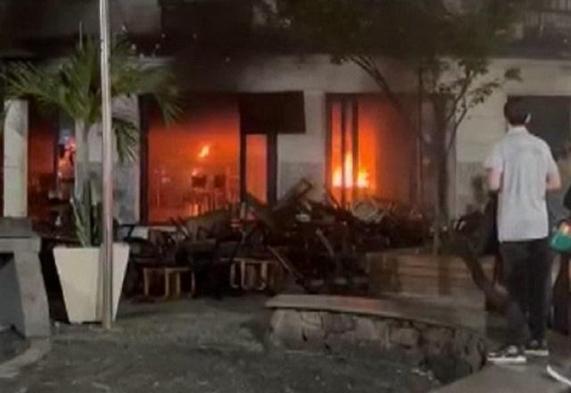 Incêndio dentro de bar durante a noite, com homem de camiseta branca em pé observando de longe
