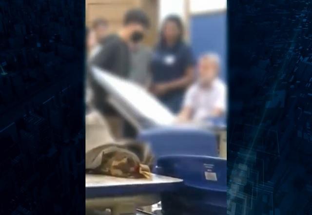 Jovem branco, de blusa preta, levantando mesa branca dentro de sala de aula, em cena de ameaça a professor, que está sentado