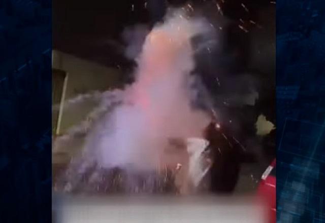 Jovem, de costas e cabelos compridos, com o braço esticado e segurando um rojão, no momento da explosão do artefato, na rua durante a noite
