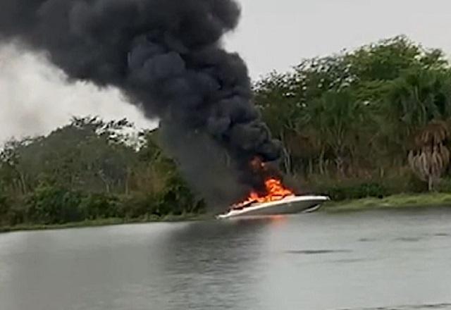 Lancha pegando fogo e soltando fumaça espessa preta após explosão em lago, com mata ao fundo