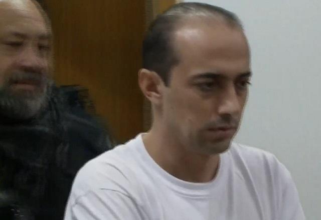Leandro Boldrini, homem branco de camiseta branca, sendo escoltado por policial fardado de barba para julgamento