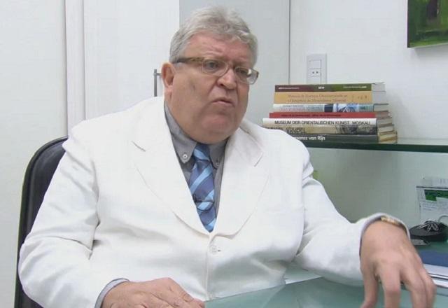 Médico, homem idoso branco, de jaleco branco, com o braço apoiado em mesa de consultório