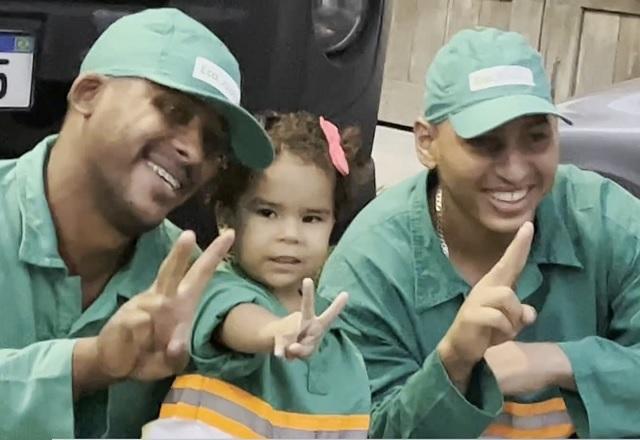 Menina de tres anos ao lado de dois garis, todos vestindo boné e uniforme verdes, fazendo sinal de V com os dedos