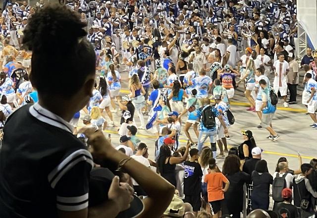 Menina negra apoiada no pescoço de pessoa de boné, olhando pessoas desfilando em passarela de desfile de escola de samba