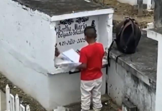 Menino negro, de camiseta vermelha, deixando papeis e flores em sepultura pintada de branco, dentro de cemitério