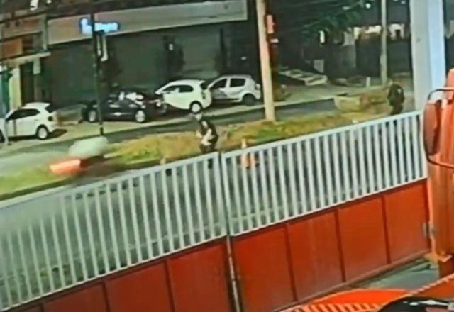 Moto se aproximando em alta velocidade, antes de atingir policial segurando cone de trânsito, em rua, durante a noite