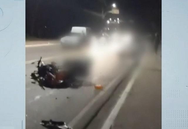 Motocicleta em asfalto, destruída, com corpos de pessoas após acidente em avenida, durante a noite