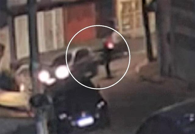 Movimentação de veículo e moto, com pessoa em pé apontando para o carro, em rua durante a noite
