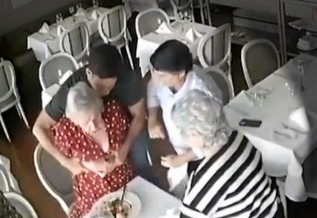 Mulher banca idosa recebendo manobra de salvamento de homem branco, ao se engasgar, sentada à mesa de restaurante