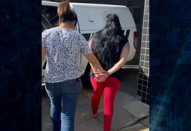 Mulher branca, de cabelos pretos e calça vermelha com as mãos para trás, algemada, sendo escoltada por policial mulher