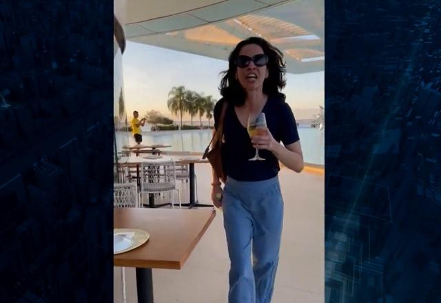 Mulher branca, de óculos escuro, caminhando dentro de restaurante com taça e garrafa nas maos, de forma agressiva