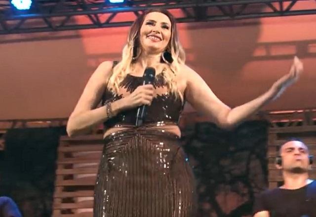 Mulher branca, loira, de vestido escuro e com os braços levantados durante show