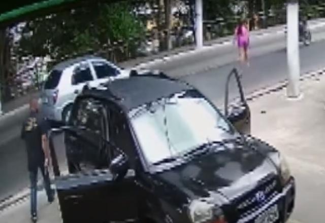 Mulher de roupa rosa correndo no meio da rua e homem de roupa preta, perto de carro, observando