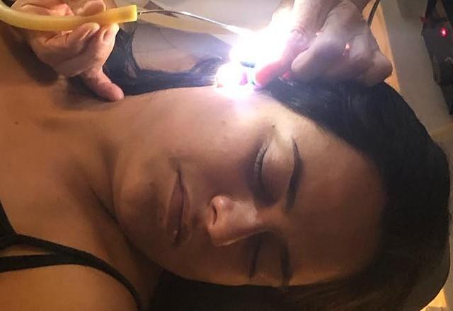 Mulher deitada em maca hospitalar com olhos fechados e médico examinando ouvido dela com uma luz