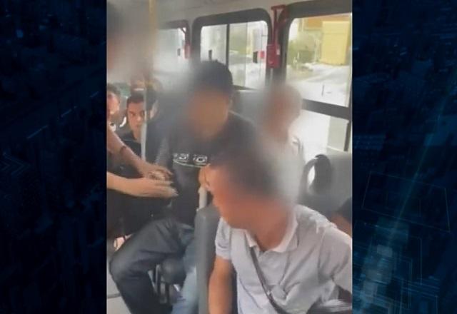 Mulher em pé apontando para celular de homem, que está sentado em poltrona de ônibus, durante o dia