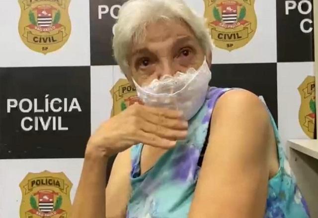 Mulher idosa, de cabelos grisalhos e máscara de proteção no rosto, em frente a painel da polícia civil