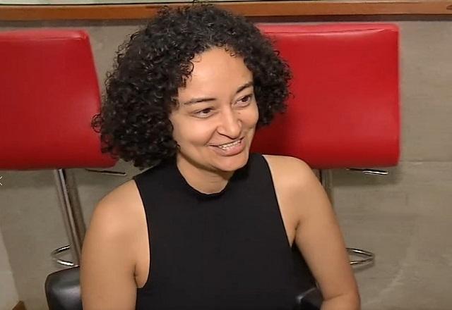 Mulher jovem, de cabelos cacheados, sorrindo durante conversa em sala, com mesa e cadeira