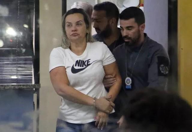 Mulher loira usando calça jeans e blusa branca sendo levada por policiais