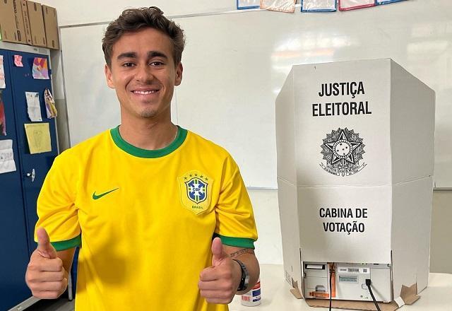 Nikolas Ferreira, com camisa da Seleção Brasileira, fazendo sinal de positivo com as mãos, próximo a cabine de votação (