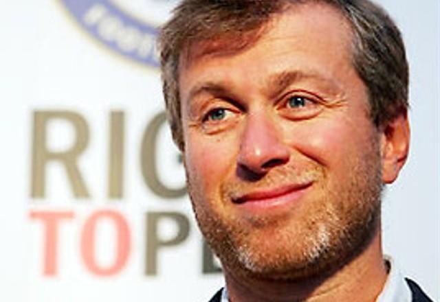 O bilionário Roman Abramovich; ele é branco, tem os olhos azuis e é loiro. Atrás. há uma foto do Chelsea, cujo dono é Abramovich