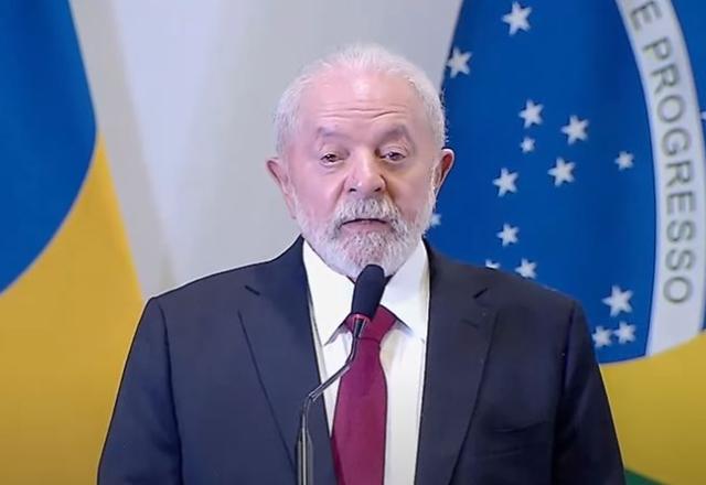 O presidente Luiz Inácio Lula da Silva (PT) de terno azul diante de bandeiras do Brasil. Reprodução