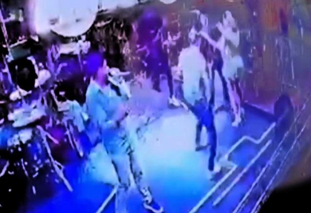 Palco de show em bar com cantor segurando microfone e, ao fundo, homem se envolvendo em confusão com mulher