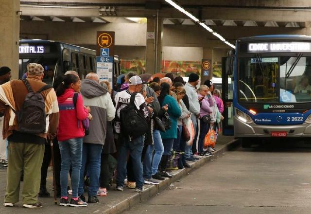 Passageiros estão organizados em fila, ao lado de um ônibus, em um terminal