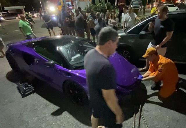 Pessoas ao lado de carro de luxo, de cor roxa, durante a noite após colisão em avenida