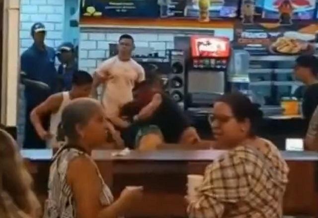 Pessoas em praça de alimentação, com homens brigando atrás de balcão de restaurante