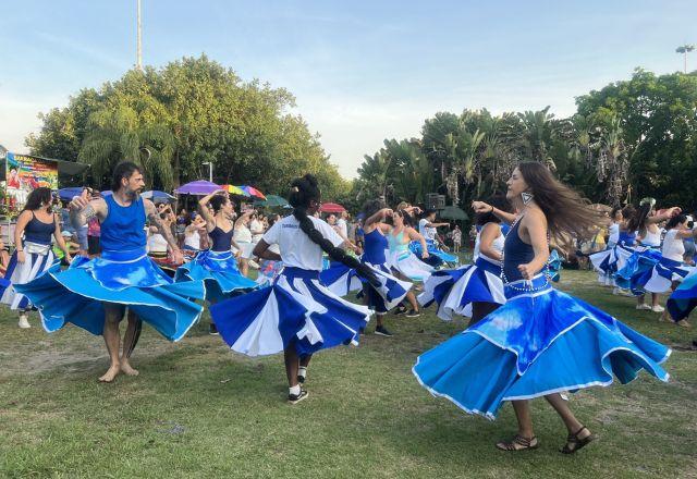 Pessoas ensaiando para o Carnaval com saias rodadas nas cores azul e branco numa área aberta e com vegetação verde
