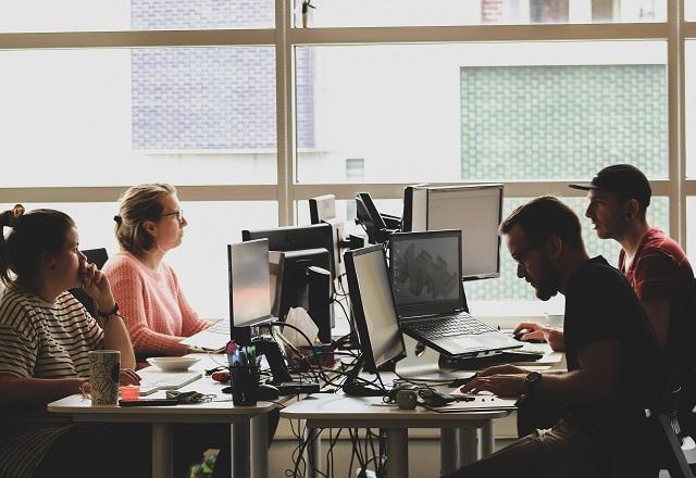 Pessoas sentadas em cadeiras, em frente a mesas com computadores, em ambiente de trabalho próximo a janela, durante o dia