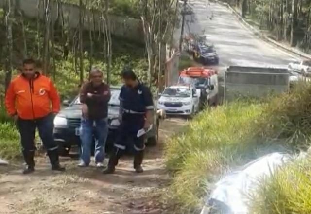 Policiais e equipes de investigação andando em área de mata, com estrada de terra ao fundo e corpo coberto por lençol branco
