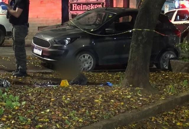 Policiais em praça, durante a noite, em frente a veículo e cabeça decapitada, com imagem desfocada
