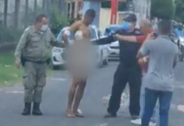 Policiais escoltando homem seminu, no meio da rua, durante o dia