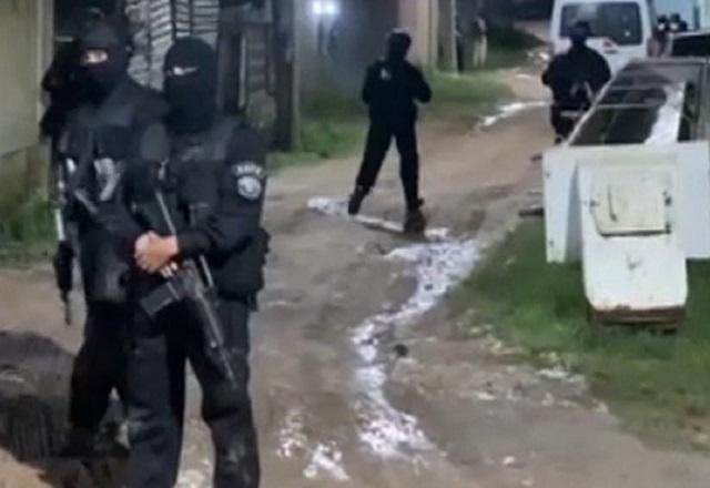 Policiais militares fardados de preto, segurando fuzis, em área aberta, durante a noite