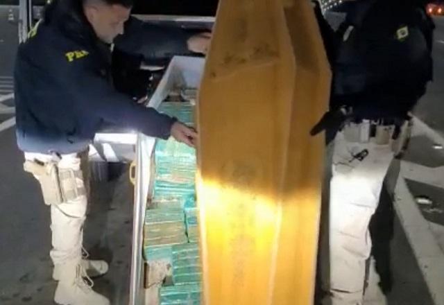 Policiais rodoviários de farda escura tirando caixão recheado com tabletes de drogas de dentro de porta-malas de carro