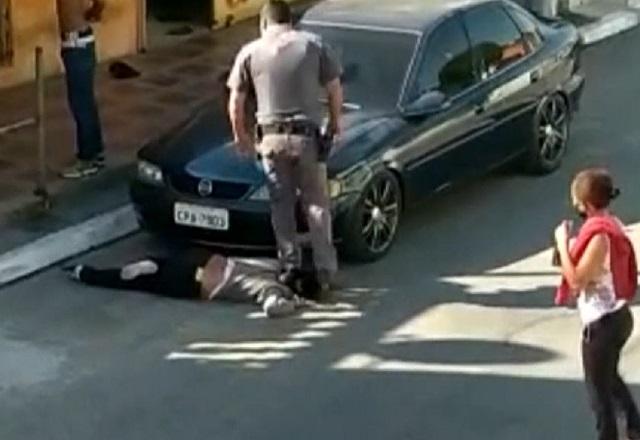 Policial fardado pisando, em pé, no pescoço de mulher negra, caída no asfalto