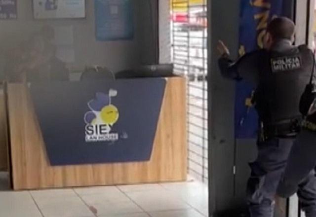 Policial fardado tentando negociar com criminoso, ao fundo e atrás de balcão, que faz mulher de refém
